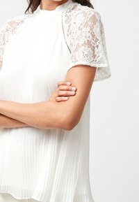 Biała bluzka z plisowanym materiałem, koronkowymi krótkimi rękawami, wysokim kołnierzem i luźnym krojem, podkreślająca mieszankę faktur i elegancji.