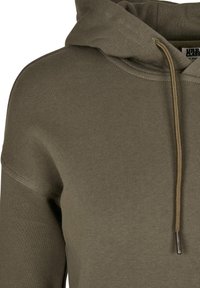 Gros plan sur le tissu d'un sweat à capuche vert olive avec des coutures visibles, un cordon et une partie de la capuche et de la zone de l'épaule.
