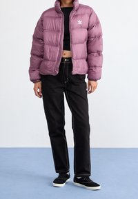 Lila pufferjacka med horisontell sömnad och hög krage, tillsammans med svarta jeans med vit sömnad och svarta sneakers.