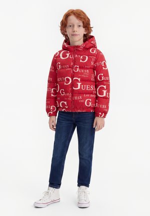 Enfant aux cheveux roux portant une veste à capuche rouge avec un texte blanc, un jean bleu et des baskets blanches, debout sur un fond blanc uni.