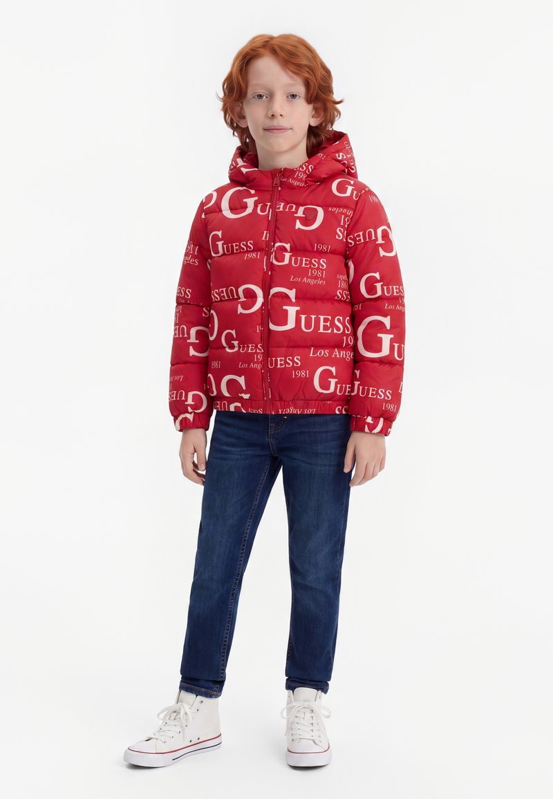 Enfant aux cheveux roux portant une veste à capuche rouge avec un texte blanc, un jean bleu et des baskets blanches, debout sur un fond blanc uni.