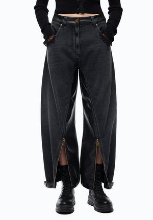 Persona che indossa pantaloni neri a gamba larga con cerniere frontali parzialmente aperte che rivelano pannelli interni effetto pelle e stivaletti neri con lacci.