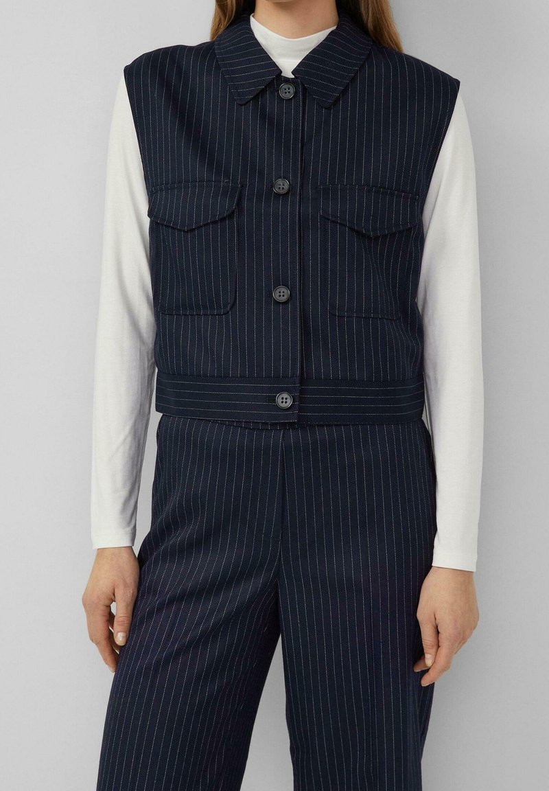 Femme portant un gilet bleu marine à fines rayures, boutonné, avec deux poches à rabat par-dessus une chemise blanche à manches longues et un pantalon assorti à fines rayures.