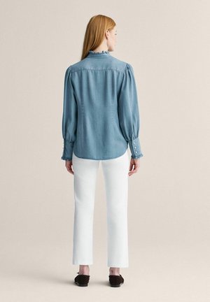 Pitkätukkainen nainen käyttää vaaleaa farkkupaitaa, jossa on pussimaiset hihat, ja valkoisia suoria housuja, seisten yksinkertaista beigeä taustaa vasten.