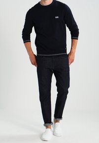 Muž v černé košili s dlouhým rukávem a malým logem, tmavých manžestrových kalhotách na značku a bílých slip-on teniskách, stojící na jednobarevném pozadí.