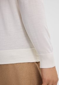 Lichtbeige gebreide trui met een geribbelde manchette en zoom, met een zachte textuur en een lichte glans, gecombineerd met een bruine kledingstuk eronder.