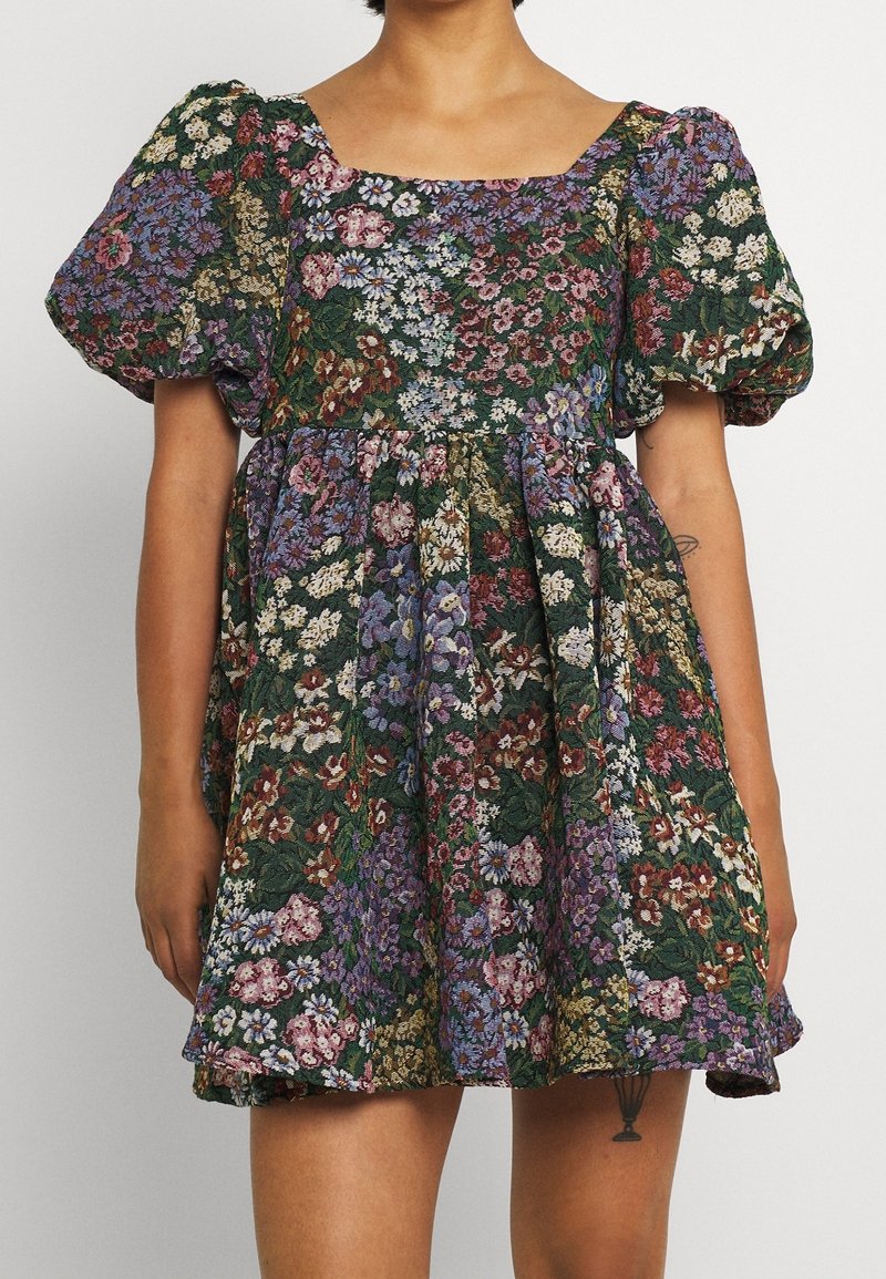 Femme portant une robe courte à fleurs avec des manches bouffantes aux couleurs vert, violet, blanc et rouge, montrant un bras et partiellement les jambes.