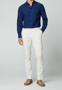 Camisa azul marino de botones, pantalones blancos y zapatos de ante marrón. La camisa tiene una tela texturizada; los pantalones son de pierna recta con un diseño limpio.