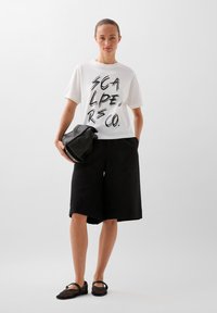 Hvid t-shirt med sort grafisk tekst, parret med løse sorte shorts. Holder en tekstureret sort clutch. Brune sko med stropper.