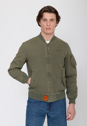 Jonge man draagt een olijfgroene bomberjack met rits aan de voorkant, zakken en blauwe jeans, staand tegen een effen lichte achtergrond.