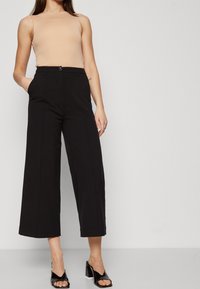 Pantalon noir à jambes larges et taille haute, avec une fermeture à boutons sur le devant et des poches latérales, porté avec un débardeur beige et des mules noires à talons.