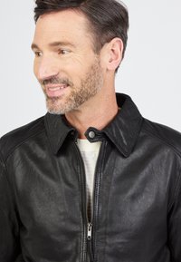 Zwarte leren jas met een rits aan de voorkant, drukknoopsluitingen en een puntige kraag. Glad textuur en getailleerd ontwerp met raglanmouwen.