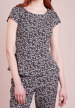 Kobieta ubrana w czarno-białą kwiecistą bluzkę z krótkim rękawem i pasujące spodnie, stojąca na jednolitym beżowym tle.