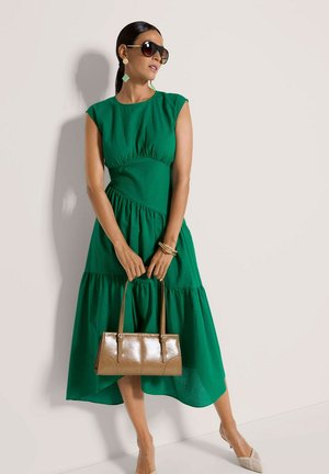 Groene jurk met textuur en een gelaagd ontwerp, mouwloze cap sleeves, gecombineerd met een glanzende bruine handtas en lichtgekleurde schoenen. Accessoires omvatten grote zonnebrillen.