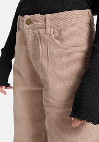 Licht beige corduroy broek met verticale ribbing, voorzien van een voorzak en een gouden knop. Zwarte ribbed lange mouwen top wordt ernaast getoond.