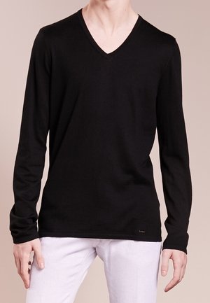 Mand iført en ensfarvet sort langærmet V-hals shirt og hvide bukser, stående mod en neutral beige baggrund.