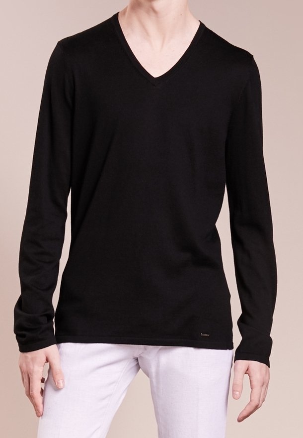 Homme portant un t-shirt noir uni à manches longues et en col en V, pantalon blanc, debout devant un fond beige neutre.
