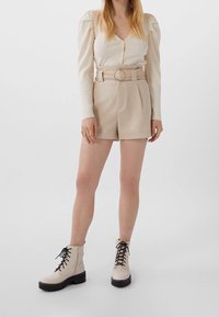 Beige geripptes Langarmoberteil mit Knöpfen, kombiniert mit hochgeschnittenen beigen Shorts, die mit einem Gürtel mit silbernem Ring versehen sind. Weiße Schnürstiefeletten.