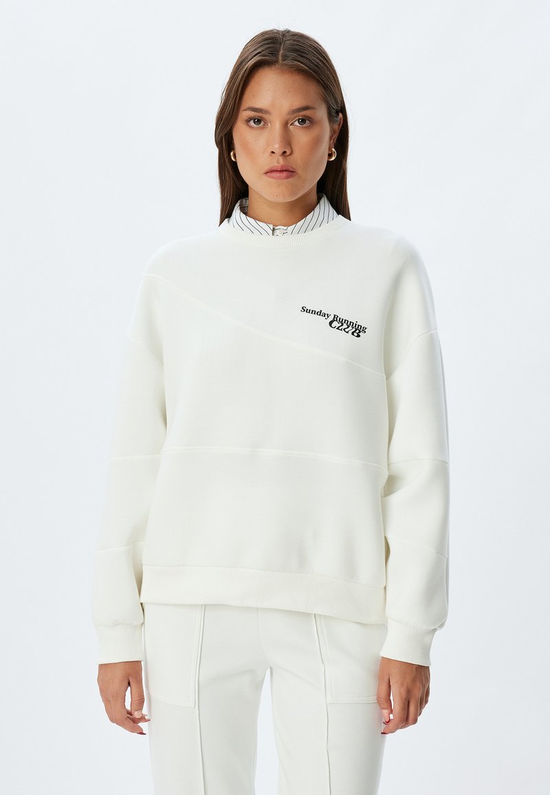 Maglione bianco con orlo e polsini a coste, caratterizzato da dettagli della cucitura diagonale e dalla scritta "Sunday Running" in nero. Indossato sopra una camicia a righe con colletto.
