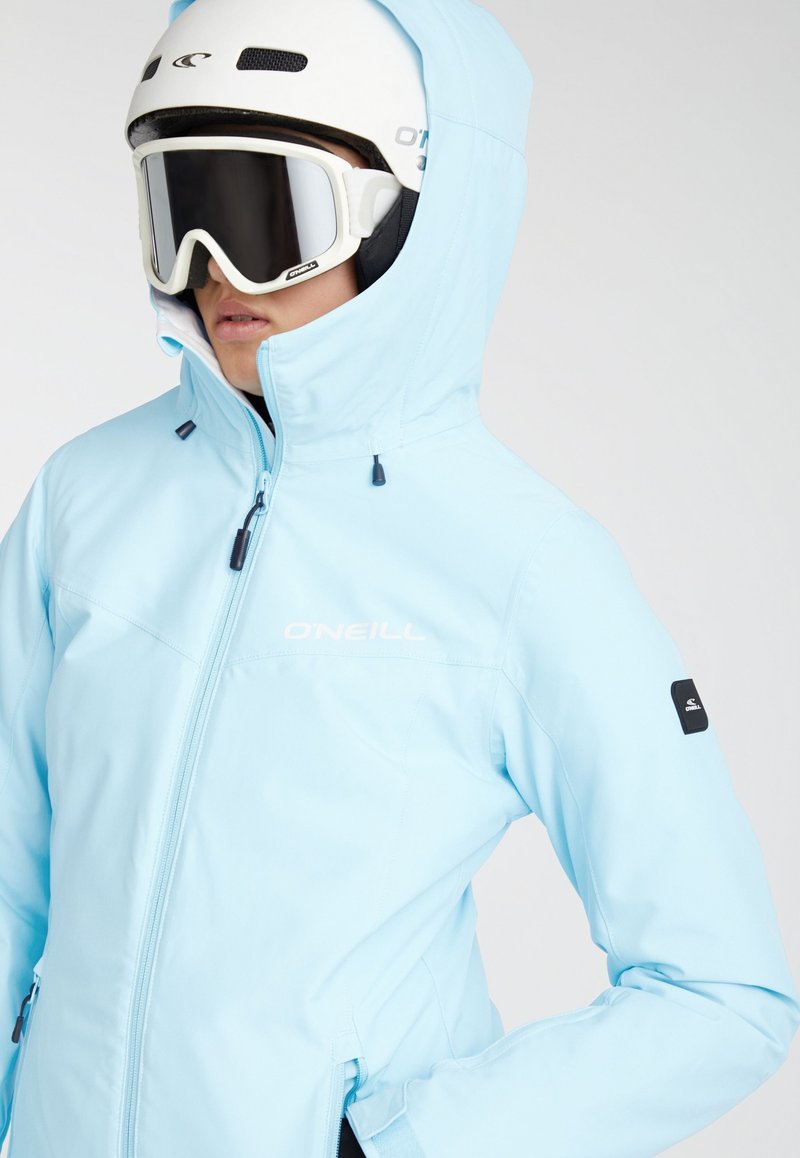 Persona che indossa una giacca da sci O'Neill azzurro chiaro con cappuccio, un casco da sci bianco e occhiali protettivi su uno sfondo neutro.