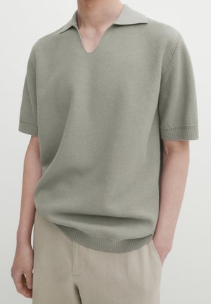 Person trägt ein hellolivgrünes, kurzärmeliges, gestricktes Poloshirt mit offenem Kragen und beige Hose, steht mit einer Hand in der Tasche.