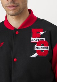Giacca varsity nera con dettagli rossi, dotata di logo e lettere ricamate del Bayern Monaco, chiusura a scatto e colletto a costine.