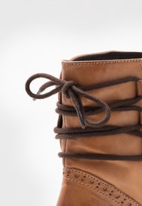 Hnědé kožené kotníkové boty s kulatou špičkou, tmavě hnědé tkané šněrování, dekorativní perforace podél svršku a hladká textura.