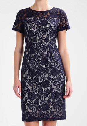 Femme portant une robe en dentelle bleu marine à manches courtes avec des motifs floraux paisley sur une doublure beige.