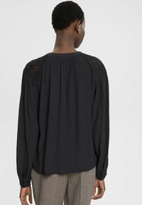 Sort bluse med lange ærmer, der har blonde detaljer på skuldrene, flydende tekstur og en samlet bagside design. Glat stof, afslappet pasform.