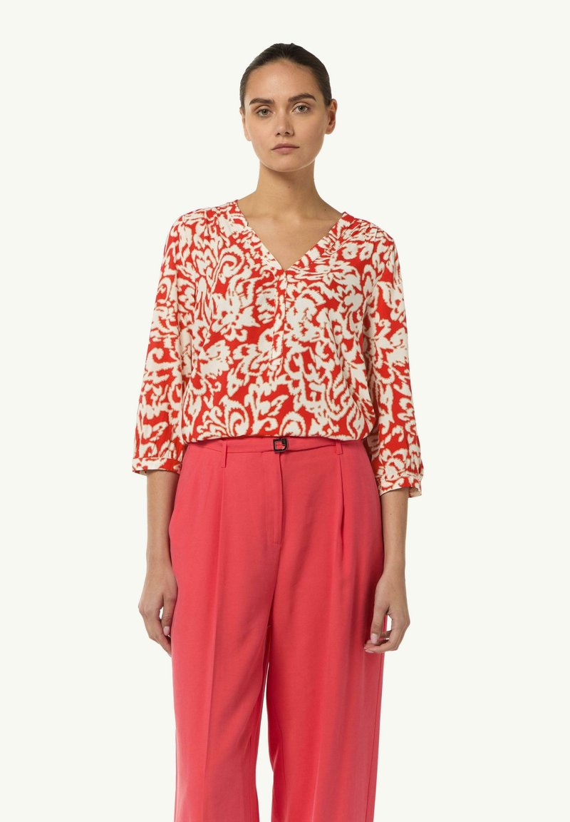 Czerwono-biała bluzka w kwiatowy wzór z dekoltem w serek i rękawami 3/4, zestawiona z różowymi spodniami o wysokim stanie. Gładka tekstura materiału.