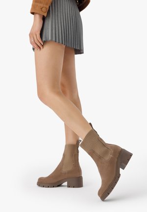 Femme portant une mini-jupe plissée grise et des bottines en daim marron à talons blocs, debout avec une jambe croisée derrière l'autre.