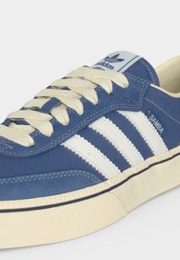 Μπλε αθλητικό παπούτσι Adidas Samba με λευκές ρίγες, κρεμ κορδόνια και κρεμ σόλα, εμφανιζόμενο από γωνιακή κοντινή λήψη.