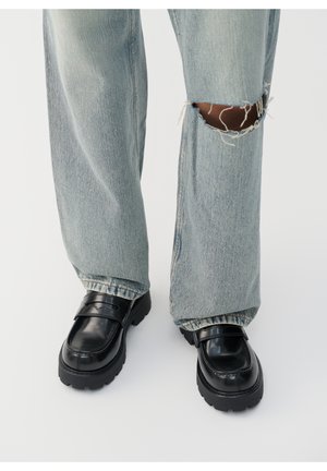 Černé lakované mokasíny s robustní podrážkou, spárované se světle modrými zesláblými džínami se znatelným otvorem na koleni.