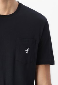 Camiseta de algodón negra con un pequeño pájaro bordado en blanco en el bolsillo del pecho izquierdo. Mangas cortas, escote redondo y textura suave.