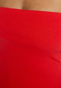 Tissu rouge côtelé avec une texture lisse, formant une forme tubulaire ajustée. Le design est sans manches et présente un col simple et épuré.