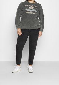 Šedá mikina s grafikou orla a nápisem "Narodil se k tomu, abych byl divoký"; černé slim kalhoty a bílé tenisky doplňují outfit. Pohodlný, uvolněný střih.