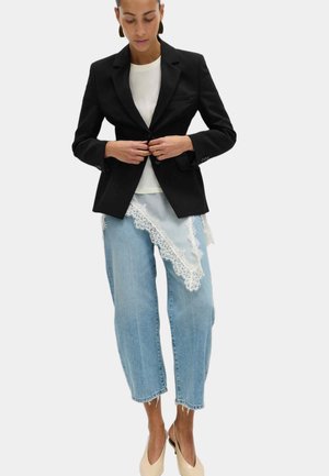 Kvinde, der knapper en sort blazer over en hvid top på, iført lyseblå cropped jeans med hvid blonde-detalje og beige slingback-hæle.