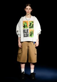 Hvid hættetrøje over en grafisk t-shirt med grønne og sorte motiver. Brune cargo-shorts med lommer. Sorte sneakers og hvide sokker.