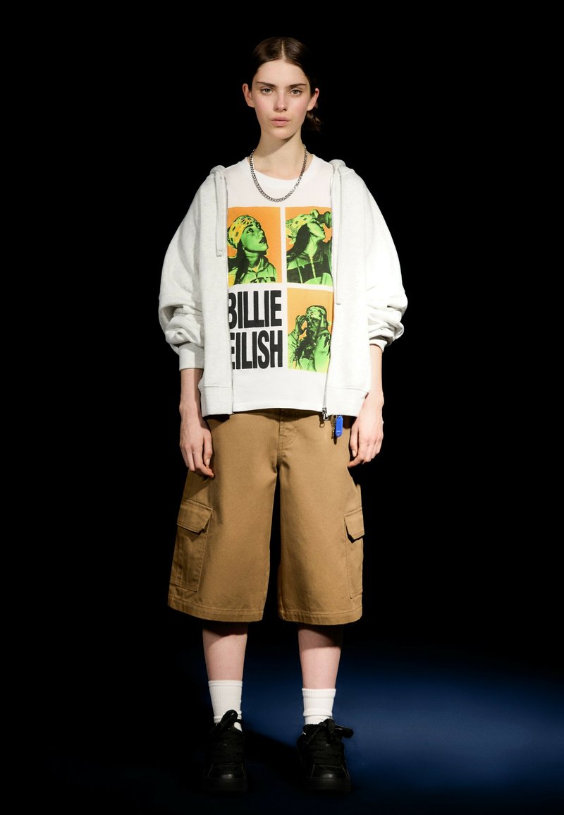 Hvid hættetrøje over en grafisk t-shirt med grønne og sorte motiver. Brune cargo-shorts med lommer. Sorte sneakers og hvide sokker.
