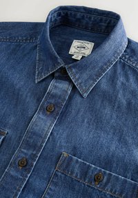 Blaues Jeanshemd mit Knopfleiste, spitzem Kragen, brauner Naht und einer einzelnen Brusttasche mit Knopfverschluss.
