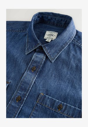 Modrá džínová košeľa na gombíky s špicatým golierom, hnedým prešívaním a jedným vreckom na hrudi s gombíkovým uzáverom.