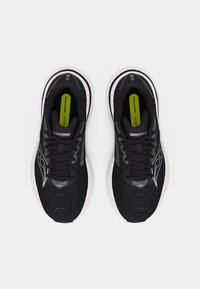 Ζευγάρι μαύρα αθλητικά παπούτσια Saucony με λευκές σόλες, εμφανές λογότυπο στις πλευρές και πράσινες εσωτερικές σόλες με την ένδειξη «Neutral», από πάνω.