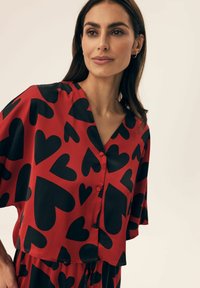 Red short-sleeve blouse with a V-neck, featuring large black heart patterns and three red buttons down the front. Smooth fabric texture.