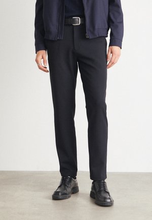 Schwarze Hosen mit geradem Schnitt, glatter Textur und klassischem Design. Kombiniert mit schwarzen Schnürschuhen und einer marineblauen Zip-Jacke.