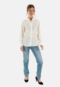 Maglietta bianca con colletto, maniche lunghe e un piccolo logo, abbinata a jeans azzurri chiari e scarpe col tacco nere.