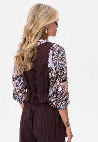 Burgundowy kamizelka w paski z koronkowym tyłem, noszona na wzorzystej bluzce w niebieskie i różowe kwiatowe wzory, z bufkami na rękawach.