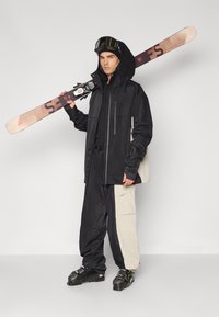 Lyžařský outfit obsahující černou voděodolnou bundu, béžové a černé kalhoty a lyžařské boty, s lyží nesenou přes rameno a brýlemi na lyže.