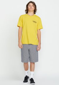 Yellow short-sleeve t-shirt with front logo, paired with gray striped shorts. Model wears black sneakers and white crew socks.