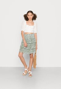 Top branco curto com mangas bufantes, detalhes em renda e cordão. Saia verde com flores em camadas. Sandálias brancas e uma bolsa de palha.