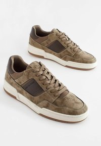 Brązowe zamszowe sneakersy z ciemnobrązowymi skórzanymi akcentami, płaskimi sznurowadłami, perforowanym czubkiem oraz białą gumową podeszwą z teksturowaną brązową podeszwą zewnętrzną.